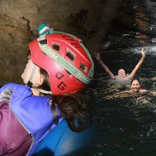 Un huésped con un casco de seguridad rojo y un chaleco salvavidas en una cámara de aire azul junto a niños nadando en una cueva de un río interior en el sur de Belice.